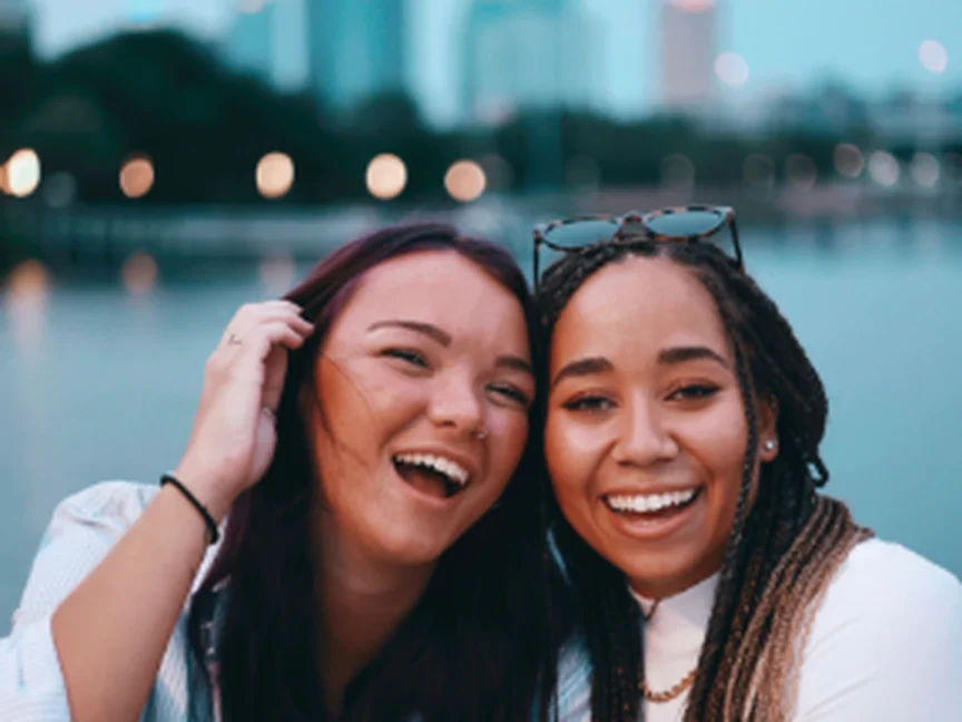 Duas jovens amigas rindo juntas à beira de um lago, com a cidade ao fundo