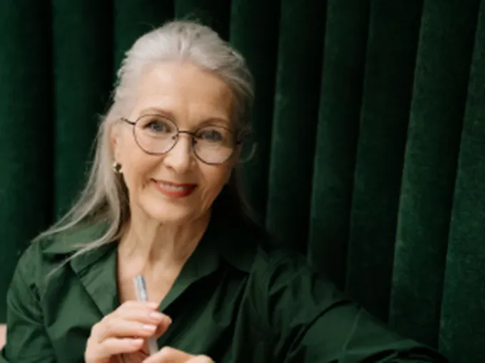 Mulher madura de cabelos grisalhos e óculos, sorrindo, sentada à mesa diante de uma cortina verde